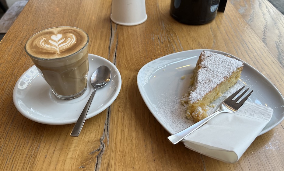 Image of a flat white with a tulip latte art in a glass and a piece of lemon ricotta cake on a white plate.