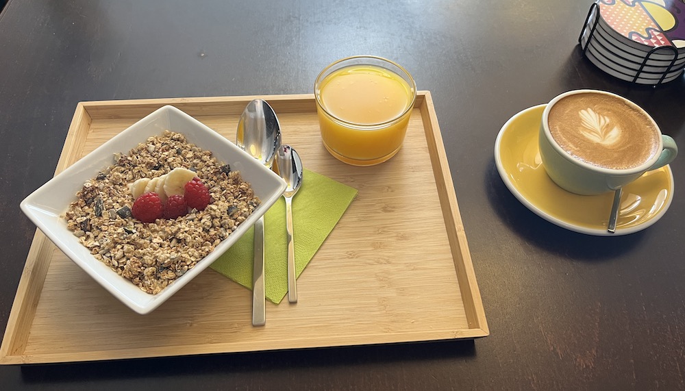 Image of a flat white with a flower latte art, a cup of fresh orange juice and selfmade granola with bananas and raspberries.