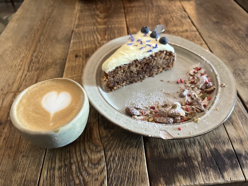 Image of a flat white with a heart latte art in a small clay cup and a piece of apple cake with a cream topping and three blue berries, as well as salted almonds on a grey plate.